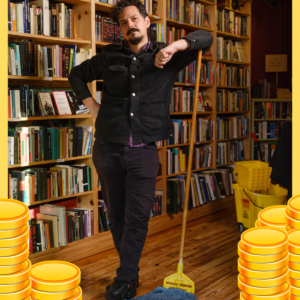 Dylan Rogers standing and leaning on a mop, in front of a bookshelf, with a border of golden coins.